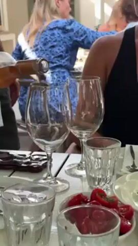 The video shows a person pouring rosé wine into two stemmed wine glasses on a white table. The table also has small clear glasses, a small bowl of halved red cherry tomatoes, plates, and silverware. In the background, a woman in a blue floral dress stands, and another person in a black tank top is seated. The setting appears to be a restaurant or outdoor dining area.