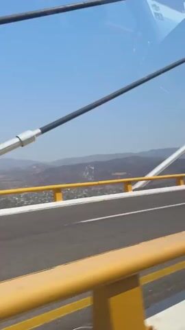The video shows the interior of a car driving across a large cable-stayed bridge on a bright, sunny day. The initial seconds (0:00-0:01) offer a view from the passenger's side window, capturing the bridge's cables, a yellow guardrail, and arid, mountainous terrain in the distance under a clear blue sky.

From 0:01, the perspective shifts to inside the vehicle, focusing on the driver from behind, the steering wheel, and the dashboard. The car's speed is visible on the dashboard display, showing 94, then 97, likely in km/h. A large infotainment screen occupies the center console, alongside two water bottles. Through the windshield, the impressive structure of the cable-stayed bridge stretches ahead, with its numerous white cables converging towards distant support towers. The surrounding landscape remains consistent with mountainous or hilly terrain.

The car's interior is light-colored, suggesting daytime travel. The audio includes sounds of people talking, laughing, and music playing, indicating a lively and positive mood among the occupants. Yellow guardrails line the roadway of the bridge. The scene is set in what appears to be a significant and scenic location, consistent with the provided context of Tula del Río, Mexico.