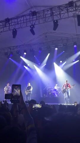 A recording from the audience shows a band performing onstage during a concert. Visible are four band members: three playing guitars and one playing drums. Many concertgoers are recording the performance with their smartphones. The stage is lit with white and blue spotlights arranged in triangular formations. The stage features an overhead metal framework rigged with lighting fixtures. The performance takes place inside a tent or temporary venue structure.