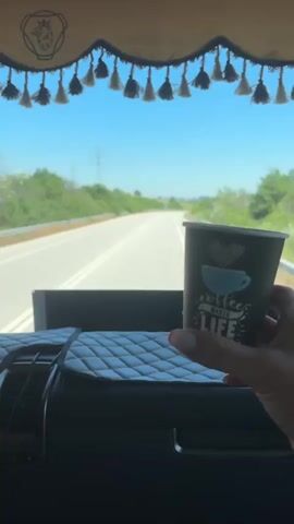 The video shows a person holding a paper coffee cup in the passenger seat of a vehicle. The cup is dark green with a white and blue design that includes a coffee cup and the words "COFFEE MAKES LIFE". The person's hand has manicured fingernails painted red and white. The background is a view from the vehicle's windshield, showing a road with green trees and a clear blue sky. The interior of the vehicle is visible in the foreground, with a quilted dashboard cover. A decorative trim with tassels hangs across the top of the windshield. The video appears to be filmed while the vehicle is in motion.