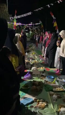 The video shows a group of people, predominantly women wearing hijabs, gathered outdoors at night. They are standing in two lines facing each other, with a long spread of food laid out on mats between them. The food appears to consist of various dishes, some in bowls and platters, with some individual portions on green banana leaves. There are also plastic bags and other food wrappers scattered on the ground. The environment is dimly lit by artificial lights, and there are decorative banners strung across the area. Some people are holding their hands up, possibly in prayer or greeting. The overall atmosphere suggests a communal meal or a religious gathering.