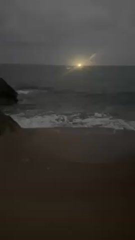 The video shows a beach with dark sand and a rocky outcropping on the left side. The sea is visible, with small waves lapping onto the shore. In the distance, there is a bright, concentrated light source that may be the sun or a bright light reflecting on the water. The sky is overcast and dark, suggesting it might be early morning or evening. There are no people visible in the scene. The camera appears to be static, providing a steady view of the scene.