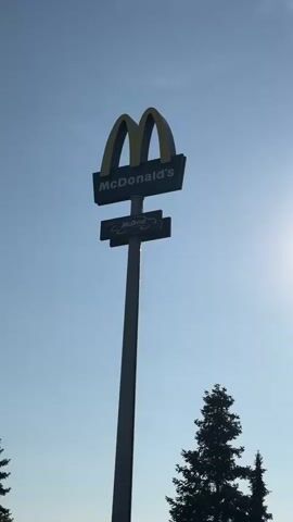 The video shows a tall sign for McDonald's against a clear blue sky. The sign features the iconic golden arches and the McDonald's name. Below the main sign, there is a smaller sign that reads "McDrive." The sun is visible in the upper right corner of the frame, casting a bright light. The silhouette of a tall evergreen tree is visible in the lower right corner, and another tree is partially visible on the left. The sign is mounted on a tall, dark gray pole. The camera angle is from below, looking up at the sign. The video appears to be a short, static shot of the McDonald's sign.