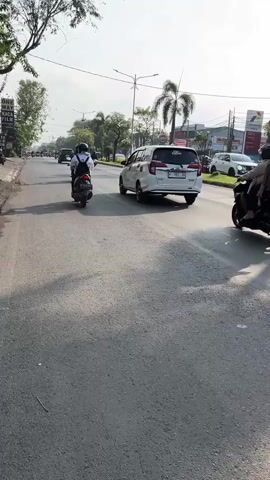The video shows a street scene with several vehicles. In the foreground, a road with a light gray asphalt surface is visible. A white van with a "Tom and Jerry" decal is parked on the right side of the road. Further down the road, a large red truck with a dark tarp covering its cargo is moving. Several motorcycles are also present, with riders in helmets. A white SUV is seen driving on the left side of the road. Palm trees and buildings, including a gas station with red and white signage, are visible in the background. The sky is bright and clear, suggesting a daytime scene.