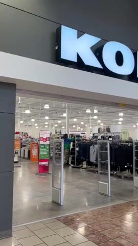 The video pans across the entrance of a Kohl's department store. A person wearing a green and blue patterned shirt and dark pants is seen inside the store, standing near clothing racks. Security gates are visible at the store entrance. To the right of the store entrance, there is a display case with items inside, and further down the mall, several other storefronts are visible, along with a red SUV parked in the mall walkway. The floor is tiled with a pattern of reddish and beige squares. The lighting appears to be typical mall fluorescent lighting.