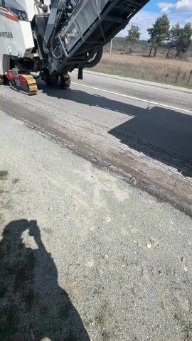 A large asphalt milling machine is seen operating on the side of a road on a sunny day. The machine, primarily white with visible "WIRTGEN" branding, is equipped with a large conveyor belt that extends over a truck. The conveyor is actively transferring asphalt debris into the bed of a white dump truck parked alongside the road. The truck has multiple axles and heavily treaded tires. The road is bordered by a gravel shoulder on one side and open fields with distant trees on the other. The sky is blue with scattered clouds. The shadow of the person recording the video is visible in the foreground.