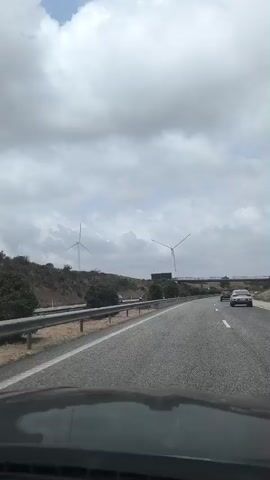 The video is taken from the perspective of someone driving on a highway. The road is asphalt with white lane markings. On the left side of the highway, there is a metal guardrail, followed by a grassy embankment with some vegetation. Two wind turbines are visible on the embankment. Further ahead, there is a bridge or overpass crossing the highway. Several cars are also visible on the highway, driving in the same direction as the camera. The sky is cloudy, indicating overcast weather. The video appears to be a short clip recorded while driving.
