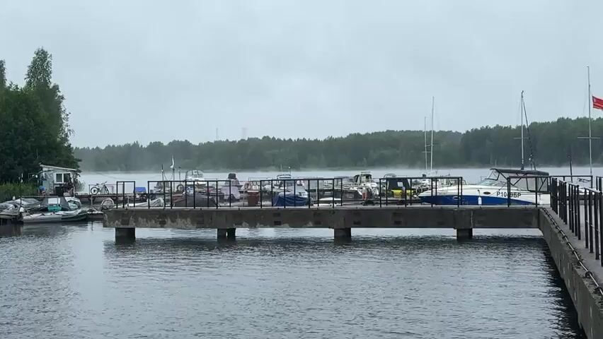 A wide shot shows a cloudy, gray day at a marina. Several boats are docked along a concrete pier. In the background, a line of trees forms the horizon. The water is calm, with a slight ripple. Mist or fog hangs low over the water, obscuring the distant trees. A small, white building with a red flag is visible on the left side of the frame. A bicycle is parked next to the building. A few people are visible on the pier, but their faces are not clear. The overall mood is overcast and still.