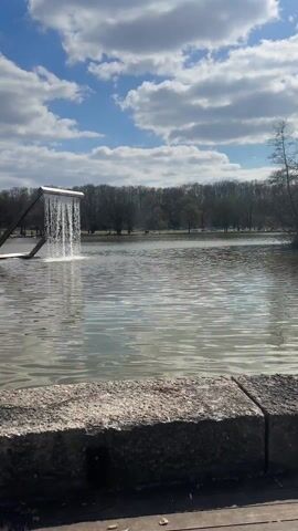 A static, medium-shot video shows a lake or pond with a rectangular waterfall-like structure in the center. The structure consists of two tilted posts supporting a horizontal beam from which water cascades into the lake. The water creates a visible splash as it hits the surface. The lake is surrounded by trees in the background, and a few people are visible walking near the lake's edge. The sky is blue with scattered white clouds. A stone ledge is visible at the bottom of the frame, partially obscuring the view of the lake.