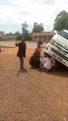 Alright, here is a matter-of-fact description of the scene in the video.

The scene shows a white van titled slightly on its side on a gravel road. The word "FAITH" is printed on the front of the van in green letters, and the license plate number appears to be KCX 782B. Four men are clustered around the van, appearing to be working on it. One man stands next to the van, another is kneeling near the front wheel, and the other two are crouching under the front of the vehicle. A fifth man walks in the background with a bicycle. The surrounding environment includes a road intersection, a building with a rusty roof, and several trees. The day appears to be overcast.