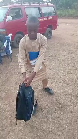 A young man is dancing in an outdoor setting. He's wearing a light brown, long-sleeved traditional outfit with a blue and white geometric design on the chest. He's also wearing black sneakers. He holds a black backpack in his hands. He spins around and moves his legs as he dances. Behind him is a red van with some text on the side, and a few chairs are visible in the background. The ground appears to be covered in dry grass and dirt. Other people are partially visible in the background, but they are out of focus. The sound suggests there is music playing.
