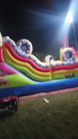 The video shows an outdoor scene at night. In the foreground, there is a large, colorful inflatable bouncy castle with a slide and circular openings. To the left of the bouncy castle, a red generator is visible on the grass. The ground is covered in green grass. In the mid-ground, there are several people sitting or standing near stalls or tents. Two scooters are parked on the grass in the right mid-ground. A large, inflated hot air balloon is visible behind the bouncy castle. The background shows more tents and lights, suggesting an event or fair. The lighting is bright, with spotlights illuminating the area. The overall impression is of a festive outdoor gathering at night.
