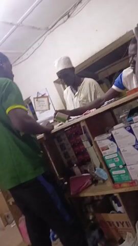 The video shows an indoor scene, likely a pharmacy or small store. There are three people visible: one customer and two people behind the counter. The customer, seen from the back, is wearing a green shirt with yellow trim and dark pants. He is at the counter, seemingly paying for an item. Behind the counter, one man is wearing a traditional white hat and a light-colored shirt, and he is handling money. The other man behind the counter is wearing a white shirt with blue sleeves, and he has one hand on his head, appearing to be resting. The counter has shelves stocked with various items, including boxes of medication, cans, and other goods. The lighting is somewhat dim, and the environment appears to be a simple retail setting.