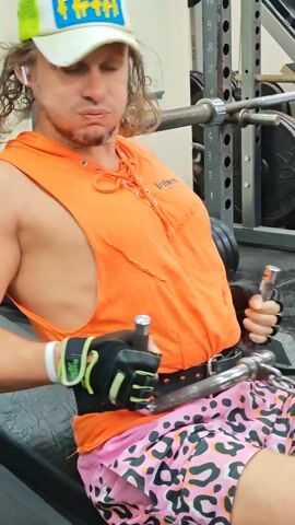 The video shows a person in a gym using a seated cable row machine. The person is male, wearing a white and yellow hat with a word partially visible on it, an orange sleeveless hoodie with lace-up details, pink animal-print shorts, a black weightlifting belt, and black and green weightlifting gloves. He has longish, wavy hair and is wearing what appears to be a wireless earbud. The gym setting includes weight plates, a squat rack, and other equipment in the background. The person is performing the rowing exercise, pulling the handles towards their stomach.