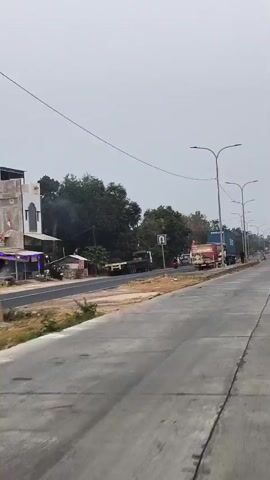 A road is visible in the video. Trees and buildings are on the side of the road. Streetlights are also visible. There are vehicles on the road. The sky is cloudy.
