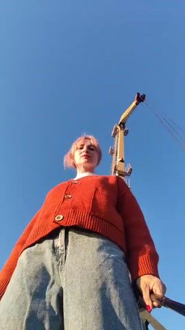 A woman with short, pink-tinted hair stands facing the camera, holding a selfie stick. She wears an orange cardigan over a white turtleneck and light-wash, wide-leg jeans. The background features a large yellow and black crane against a clear blue sky. The woman is positioned on a concrete platform, possibly a dock or industrial area, with a body of water visible to her left and piles of gravel or sand to her right. The sun appears to be low in the sky, casting a warm light.