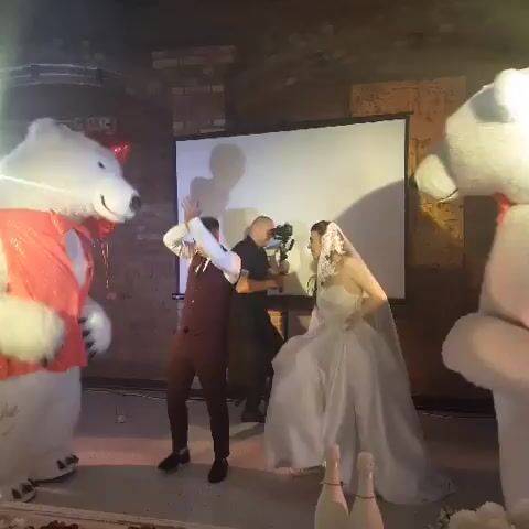 The video opens with a view of a wedding reception in a brick-walled venue. A person in a white polar bear costume, adorned with a red sequined vest and a red heart balloon, stands on the left. A man in a maroon vest and tie, and a bride in a white wedding dress and veil are center stage. Another person, dressed in black, stands behind the bride, holding a camera or video recording device. A projector screen is positioned behind the bride and groom. The polar bear costume character gestures with its hands, and the man in the maroon vest also makes hand gestures, appearing to dance. The bride looks down as she moves. The video seems to capture a moment during a wedding celebration, possibly involving entertainment or a performance.