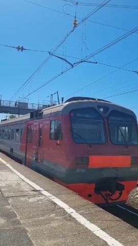 A red and gray electric train is stopped at a station under a bright blue sky. The train is positioned on the right side of the frame, with its front facing towards the viewer. A concrete platform runs along the left side of the train, with a white line marking the edge. To the left of the platform, there is a set of stairs leading up to a pedestrian bridge that spans over the train tracks. Several people are visible on the stairs and the bridge. The train has large windows at the front and appears to be a passenger train. Overhead power lines and poles are present above the train and tracks. Trees and buildings can be seen in the background.