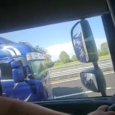 The video shows the view from inside a vehicle, looking out the window. To the left, a blue semi-truck with a white eagle logo and crown is visible. The truck has the word "SCANIA" in white text on the side. To the right, another semi-truck is passing by, also blue. The side of this truck has a large white logo that appears to be text, with red text below it. The truck is on a highway, with other vehicles visible in the distance. The sky is clear and blue.