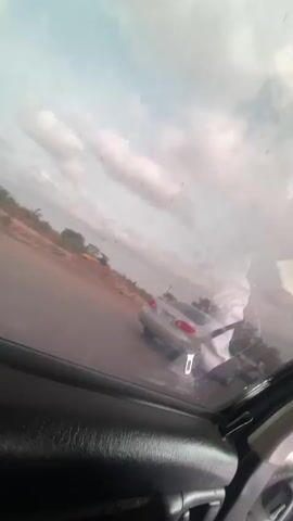 The video is taken from inside a moving car, looking out the window. Initially, a silver car is visible in the distance. As the car filming moves forward, a large truck covered with a blue tarp passes by. The truck has green and red elements, and the red wheels are visible. The sky is cloudy.