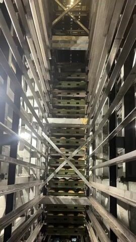 The video shows a vertical shaft with a series of stacked shelves or platforms. Each shelf appears to hold multiple round, light-colored objects, possibly loaves of bread or rolls. The structure is made of metal, with beams and supports forming a cage-like enclosure around the shaft. The perspective is looking straight down the shaft from above, giving a sense of depth and height. The lighting is dim, with some light sources visible on the left side of the frame, casting shadows and highlighting the metal structures. The overall impression is of an industrial or storage environment. There are no people or overt actions visible in the video.