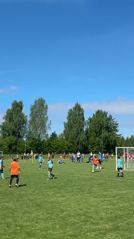 The video shows a children's soccer game on a green field with trees and a bright blue sky in the background. Players in orange and blue jerseys are scattered across the field, actively playing. A referee in a yellow vest is overseeing the game. There are goals at both ends of the field, and spectators can be seen along the sidelines. Some children are sitting on the ground, presumably watching or resting. The overall atmosphere is one of energetic activity and sunny outdoor sports.