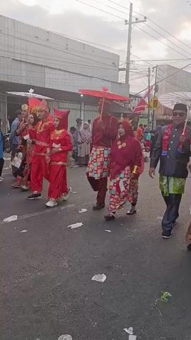 A group of people are walking down a street. Some are wearing traditional clothing and carrying umbrellas. There are onlookers on the side of the street. There are buildings and power lines visible in the background. The ground is littered with trash. One person is holding a small Indonesian flag.