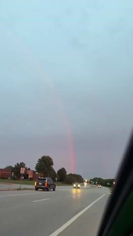 A car is driving down a highway with a rainbow in the sky. The sky is cloudy and the sun is setting. The rainbow is visible in the upper left part of the sky. The car is in the right lane and is moving away from the camera. There are other cars on the road. The environment is an urban or suburban setting with trees and buildings visible in the background.