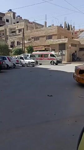 The video depicts a street scene in a Middle Eastern town. The primary subject is a white and red ambulance with Arabic writing on the side, labeled "AMBULANCE" in English. The ambulance has a small trailer attached. It is parked on a street with several other cars. Buildings in the background appear to be older, with some damage visible on the facades. A sign in Arabic is also visible on one of the buildings. A person is standing near the building with the sign. The street is paved and shows signs of wear.