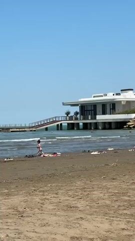 A young child wearing a pink swimsuit is running near the water's edge on a sandy beach. There are several pieces of clothing scattered on the sand in the foreground. In the background, a modern white building with a flat roof is situated on stilts over the water, accessed by a ramp. The sky is clear and blue, and the water appears choppy.