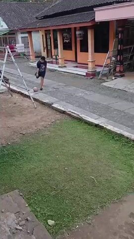 A young boy in an orange shirt and blue shorts is playing with a ball in a paved courtyard in front of some buildings. He kicks the ball and then runs after it. The buildings appear to be houses with a scooter parked in front of one. There is grass and a concrete structure in the foreground.