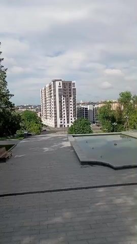 This media file captures a panoramic view of an urban landscape in Kharkiv, Ukraine. The foreground features a wide, gray paved area, possibly a plaza or a park pathway, with a simple wooden bench visible on the left. Adjacent to this is a large, rectangular pool or fountain with water reflecting the sky.

Dominating the midground is a tall, modern apartment building with a white facade and darker accents, featuring numerous balconies and windows. Surrounding the building and extending to the background are lush green trees, indicative of a park or a tree-lined urban area. Further in the distance, other buildings, including what appears to be a lower-rise modern structure, are visible, forming part of the city's skyline. The sky is partly cloudy, with fluffy white clouds scattered across a pale blue expanse.

The scene suggests a tranquil daytime setting, likely late morning or early afternoon, given the bright lighting and the shadows cast on the paved area. The weather appears pleasant, with a clear sky and visible greenery. There are no people clearly visible in the frame, lending a sense of quietude to the scene. The overall impression is one of a well-maintained public space integrated within a developing urban environment. No text is discernible in the media file.