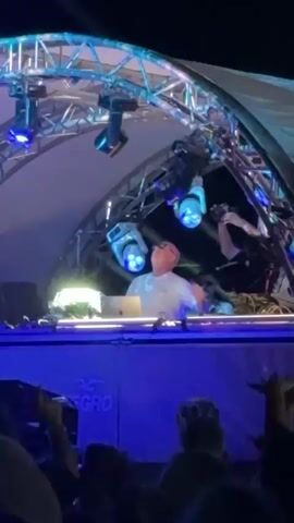 A video of a DJ performing at an outdoor concert. The DJ, a bald man wearing glasses and a white t-shirt, stands behind a table with a laptop and DJ equipment. He is illuminated by stage lights, which flash and change color. A crowd of people are visible in the foreground, with their hands raised in the air. The concert appears to be taking place at night, with a dark sky and a large structure resembling a sculpture in the background.
