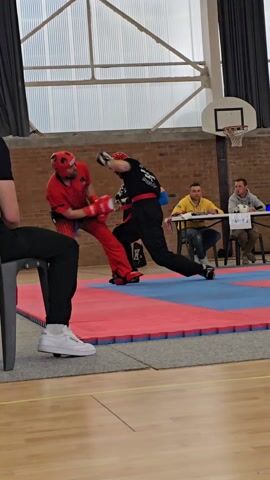 A martial arts sparring match is taking place. A person in red protective gear faces off against a person in black, who attempts a kick towards the other, with both wearing headgear and gloves. There are three people seated at a table on the side watching the spar. One person sits to the left, partially obscured, and a photographer stands in the background. The environment looks like an indoor gymnasium or arena with a basketball hoop visible.
