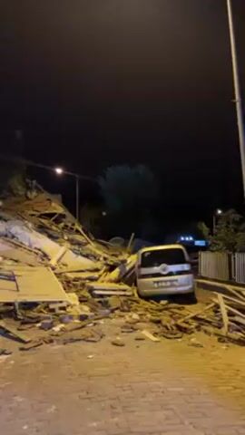 At night, the scene reveals the extensive destruction of a building, collapsed into a pile of debris. The wreckage is scattered across the ground, composed of building materials like concrete, wood, and metal. Adjacent to the debris lies a silver car parked on a brick-paved road. Streetlights illuminate the scene, highlighting the severity of the collapse and its impact on the surrounding area. The night sky is dark, and there are trees visible in the background.
