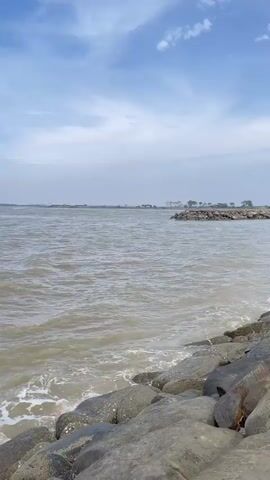The video shows a view of a large body of water, likely a sea or ocean, from a rocky shore. The water is choppy with small waves breaking against the rocks in the foreground. The sky is partly cloudy with a mix of blue and white. In the distance, a hazy shoreline is visible with some structures or buildings. The overall scene depicts a natural coastal environment.