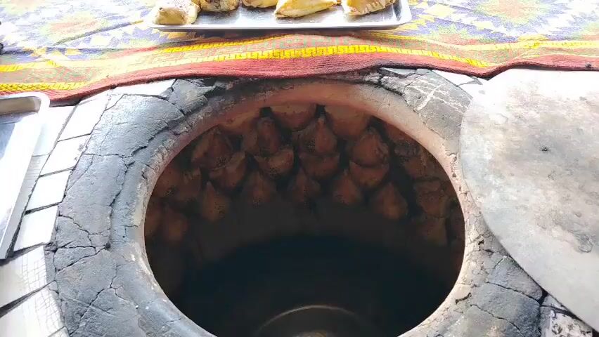 This media file captures a close-up view of a tandoor, a type of clay oven, filled with freshly baked bread. The bread, characterized by its golden-brown crust and a dusting of sesame seeds, is arranged in rows along the interior walls of the oven. The tandoor itself appears to be constructed from rough, grey clay, showing some signs of wear and cracking around its opening. A decorative, colorful fabric with red and yellow patterns is visible at the top, framing the oven. The scene is lit with natural light, suggesting it is daytime. The focus is entirely on the bread and the tandoor, with no people or further environmental details visible. The activity is clearly the baking of bread, a common culinary practice in Central Asia, including Kyrgyzstan, where Ala-Buka is located. The visual suggests a rustic, traditional setting, emphasizing the handmade nature of the food. The close proximity of the shots to the bread highlights its texture and the process of baking.