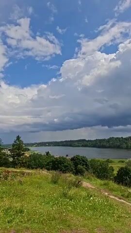 A scenic overview shows a vast lake bordered by lush green trees and vegetation. The sky above is a mix of blue patches and large, fluffy white clouds, with some darker, more ominous clouds in the distance. A dirt path winds through the grassy foreground, suggesting a natural, outdoor setting. The overall impression is one of a serene, perhaps slightly moody, summer landscape.