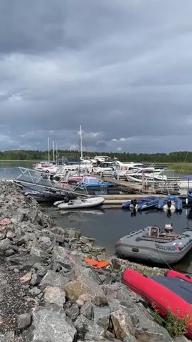 The video shows a marina with numerous boats docked. In the foreground, there are several inflatable boats, some with outboard motors. In the middle ground, a wooden pier juts out into the water, lined with a variety of boats, including speedboats and a sailboat. In the background, more boats are visible, along with a tree-lined shore and a cloudy sky. The water appears calm, and the overall atmosphere is serene.