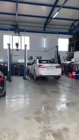 In a well-lit auto repair shop, a white sedan with a license plate that reads AE 7490 IA is elevated on a two-post lift. The car is positioned in the center of the shop, facing away from the camera. The floor is made of concrete with a slight shine and some visible wear. To the left of the car, there is a dark-colored vehicle parked, partially visible. The shop contains standard equipment for auto repairs, including tool cabinets, a chain hoist hanging from the ceiling, and stacked tires in the background. Natural light enters through multiple windows at the back of the shop, and artificial lighting is provided by overhead fluorescent lights.