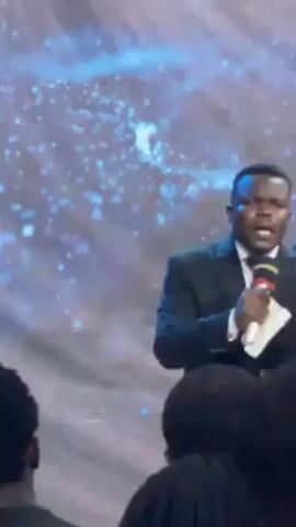 A man wearing a suit and tie speaks into a microphone in front of a backdrop with a blue and white pattern suggesting stars. He is looking at the audience and appears to be emphasizing a point. There are people in front of him, but their faces are not fully visible.