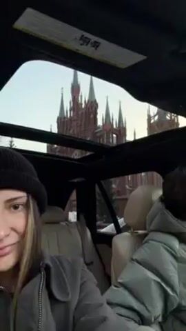 Two people are in the back seat of a car, looking up through the open sunroof at a large, red brick Gothic Revival-style church with spires, likely in Russia. The person on the left, a young woman with blonde hair and a black beanie, looks at the camera and smiles slightly. She's wearing a gray jacket. The other person is mostly out of frame. The interior of the car has light-colored leather seats. The sky is overcast.