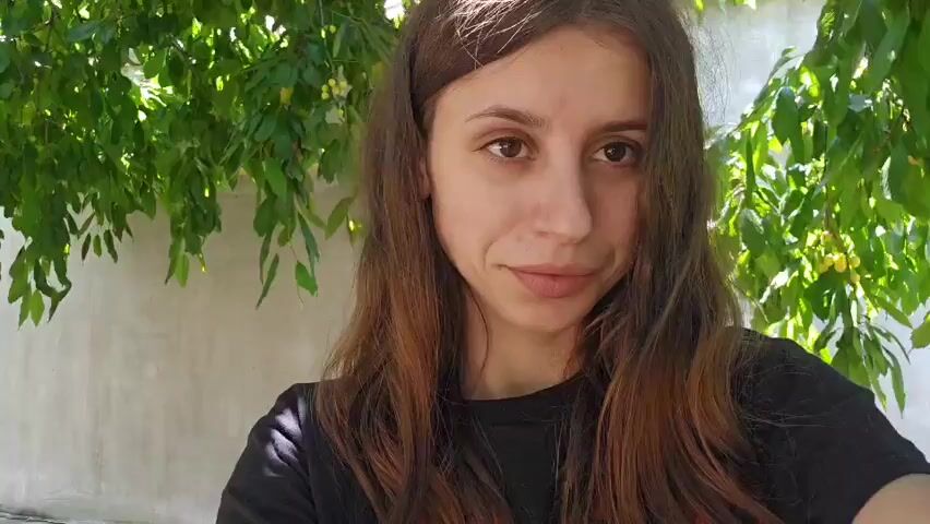 The video begins with a close-up selfie of a young woman. She has long, wavy brown hair and is wearing a black t-shirt. She is outdoors, with green foliage visible in the background, suggesting a garden or park setting. The woman smiles faintly at the camera. The video then transitions to an extreme close-up of her eyes. Her right eye is looking slightly to the left, while her left eye looks directly at the camera. The focus is sharp on her irises and eyelashes. The background details are blurred, further emphasizing her facial features.