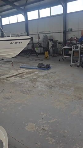 The video shows the interior of a large, industrial-style building with a gray concrete floor and corrugated metal ceiling. There are several white boats, each bearing names like "NISSI 8," "EVAGORAS 1," and "OTHELOS," parked within the space. The boats are either on trailers or on stands with wheels, facilitating their movement. In the background, there are various pieces of equipment, including a black object on a blue mat. Light streams in through large, rectangular windows along the top of the walls. The camera pans slowly across the scene, offering a view of the various boats and the tools within the building.