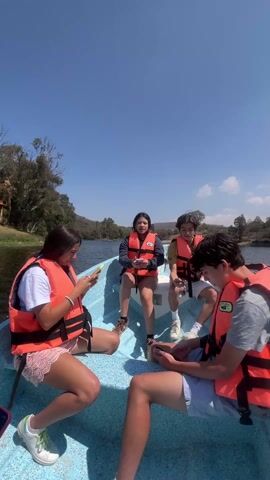 The video shows four young people on a blue motorboat in the middle of a lake. They are all wearing orange life vests. A young woman on the left is looking at her smartphone. Another woman is sitting next to her, talking. Two young men are seated on the right side of the boat, both looking down. The boat is moving, as evidenced by the wake it is creating. The sky is blue with some scattered clouds. The shoreline is visible in the background, with trees and vegetation.