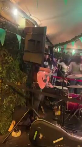 The video shows a live band performing on a small stage outdoors, in what appears to be a pub or patio area decorated with green shamrocks and string lights, likely for St. Patrick’s Day. A man plays an acoustic guitar while another man sits behind him playing the drums. The band is positioned in front of a large black speaker and a black guitar case. A crowd of people, mostly young adults, are dancing and mingling near the stage. Some individuals in the crowd are holding phones. The scene is lively and festive with people enjoying the music.