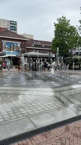 The video shows an outdoor plaza with people walking around and a water fountain feature in the foreground. The fountain consists of jets of water spraying upwards from the ground, some of which are lit with purple lights. In the background, there are buildings, including one with a McDonald's sign. Several green trees are visible, and umbrellas shade outdoor seating areas. The weather appears overcast. Some children are seen playing near the fountain.