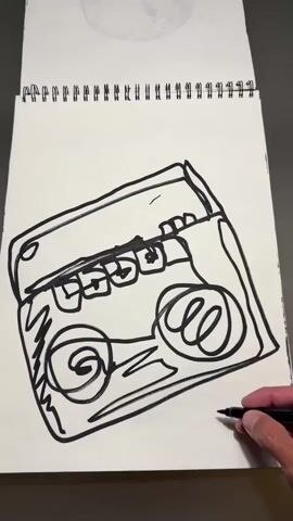 A person's hand holds a black marker and is drawing on a white spiral-bound sketchbook. The drawing is a black-line sketch of a boombox. The drawing appears to be a quick, stylized sketch with bold lines and some scribbled details. The sketchbook is open and lies flat on a dark surface. The background is out of focus but suggests a neutral environment. The drawing is approximately in the center of the sketchbook page. The hand and marker are in the lower right corner of the frame, actively engaged in adding detail to the drawing, specifically on the lower right side of the boombox, possibly signing or adding a final flourish. The drawing itself shows a rectangular body with two speaker grilles on the front, a cassette deck with buttons on top, and some antenna detail at the top. The sketch is unfinished, with the hand actively adding more lines.