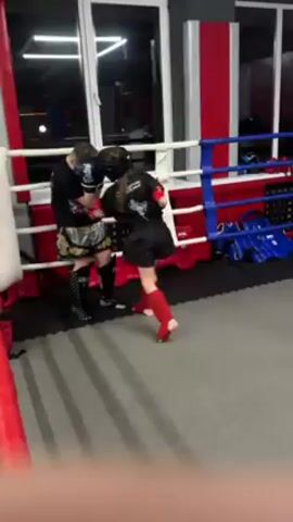 Inside a boxing ring, two young fighters engage in a sparring session. The ring has red, white, and blue cushioned corners. The first fighter is taller, wearing a black t-shirt, patterned shorts, and black sparring gloves. The second fighter, who appears shorter, is clad in a black t-shirt with a logo, matching shorts, red protective leg gear, and a head guard. They are facing each other, throwing punches and practicing defensive maneuvers. The ring's floor is covered in grey matting. In the background, one wall features a large window, while protective gear rests along one side of the ring.