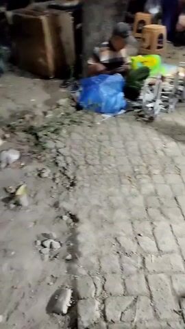 The media file captures a street scene in Sukodono, Indonesia, likely at dusk or night due to the artificial lighting and shadows. The primary subjects are people and various objects laid out on the ground.

In the foreground, a person wearing a red headscarf and patterned pants walks away from the camera. To their left, a person wearing a cap and a striped shirt is seated on the ground, surrounded by several items including a large blue plastic bag, a green object, and what appear to be small, wheeled carts. Behind them, a large cardboard box is visible, leaning against a tree. Further in the background, there are more people, some standing around what looks like a food cart with a red exterior and a white canopy. Several small, wooden stools are also present. The ground is a mix of dirt and paved brickwork, with some debris scattered around.

The overall activity appears to be a street market or a gathering where goods are being sold or displayed. The presence of the food cart suggests a place where people might be eating or socializing. The lighting and the arrangement of items indicate a commercial or informal gathering taking place outdoors. There are no overtly discernible emotions, but the scene suggests a typical evening street scene in a community setting. No visible text is clearly legible in the provided frames.